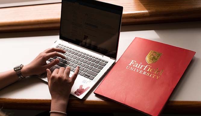 A student uses a laptop which sits next to a red folder titled “Fairfield University” in gold lettering.