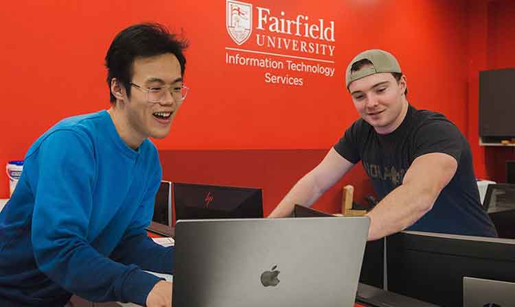 Image of two students collaborating on a laptop at the Fairfield University help desk in the library.