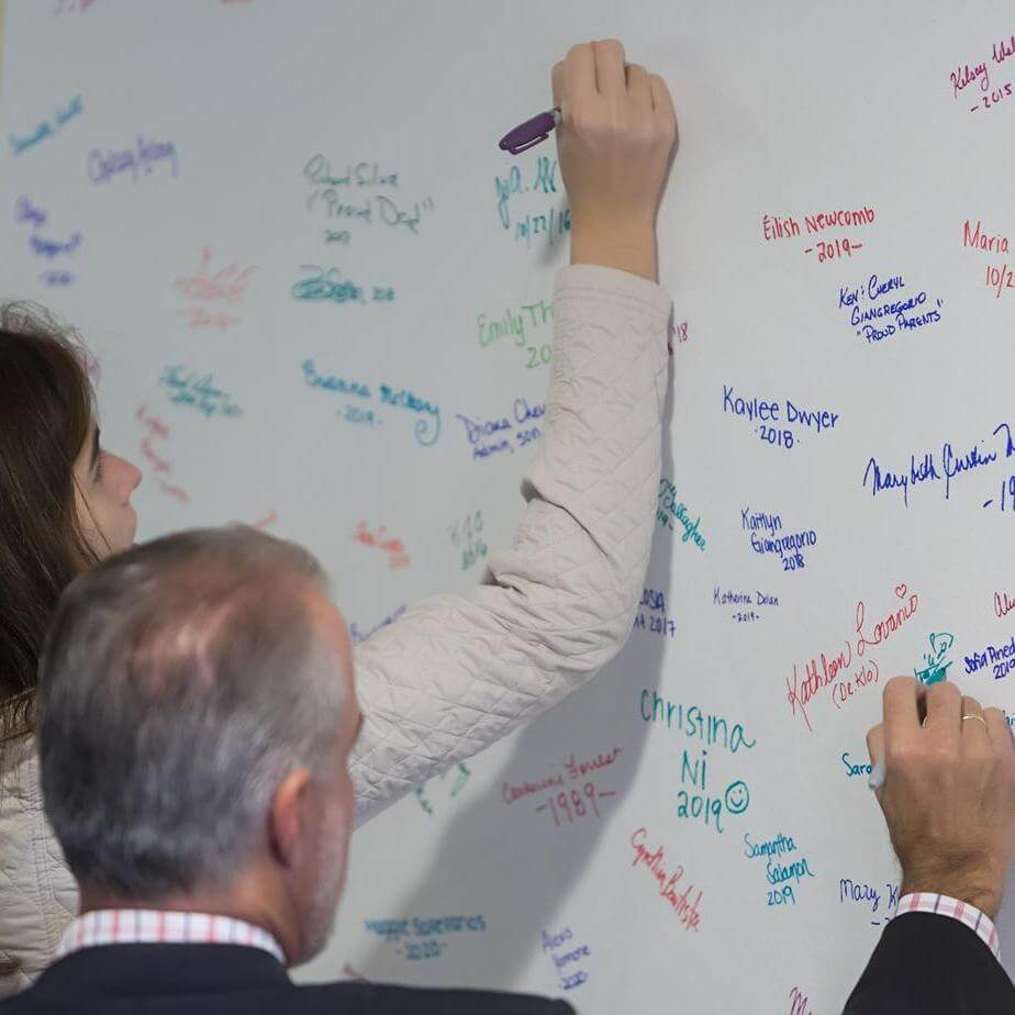 Two alumni sign their names with sharpies on a large piece of white paper. 