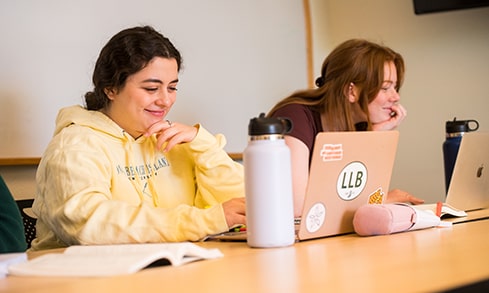 Two students in a classroom doing schoolwork with notebooks and laptops.
