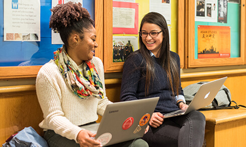 Two students are seated on a bench, each using a laptop, appearing to work together in a classroom setting.