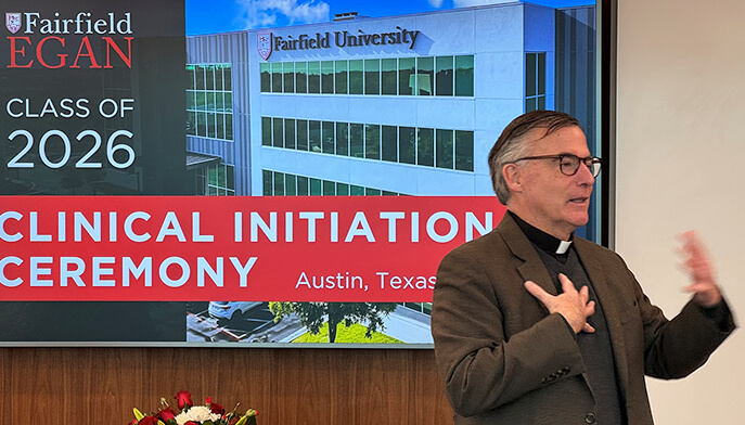 Rev. Kevin O’Brien, S.J. presents information to an audience, positioned in front of a large screen filled with graphics and text.
