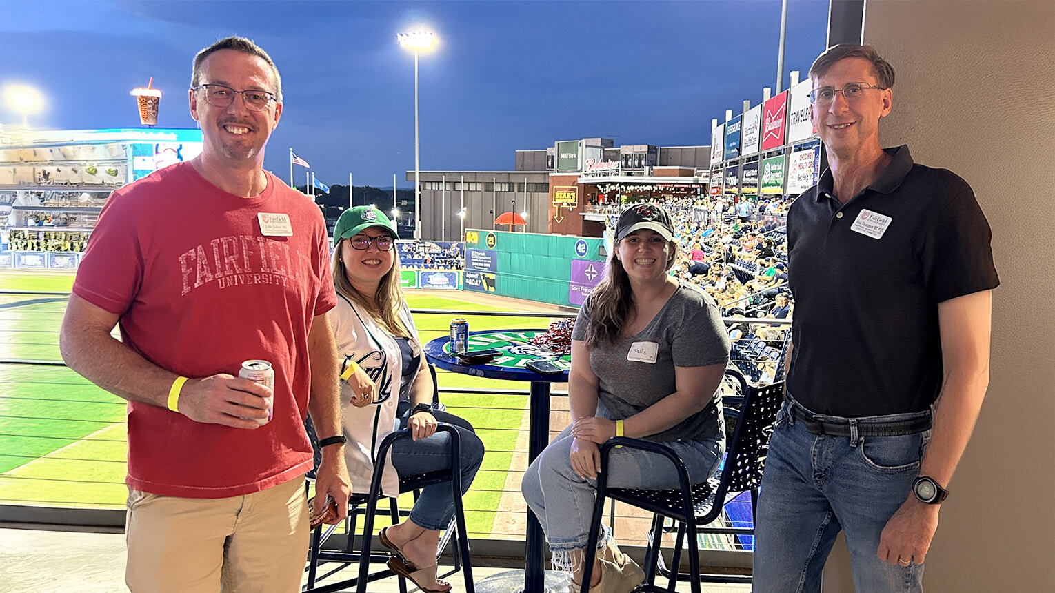 A group of four people gathered around a table, watching a baseball game in an energetic outdoor setting. 