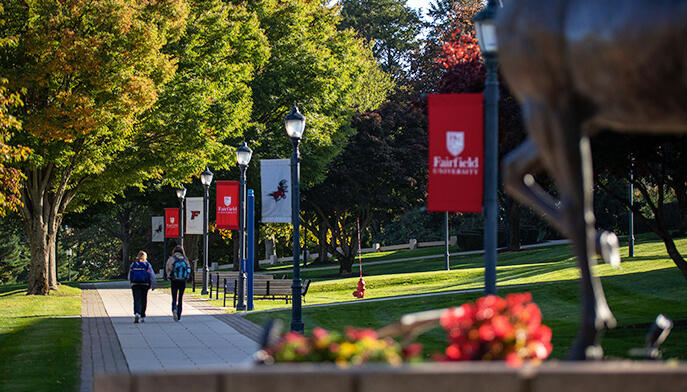 Two people walk on a path on campus next to a stag statue, with trees and grass framing the scene.