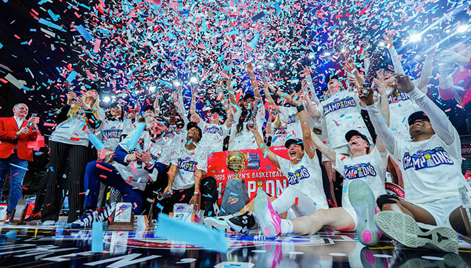 The women's basketball team joyfully celebrates their conference championship victory, embracing and cheering together. 