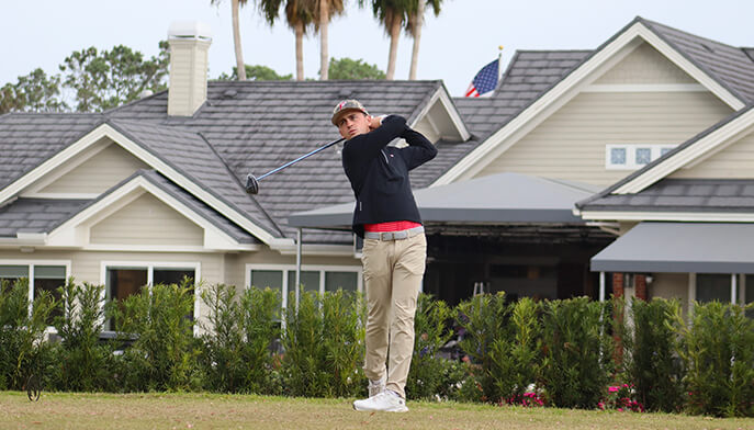 A student on the Men's golf team wearing a black shirt and khaki pants swings a golf club on a green golf course. 