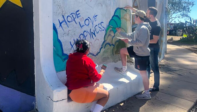 Three individuals paint a colorful graffiti mural on a large wall, showcasing their artistic collaboration.
