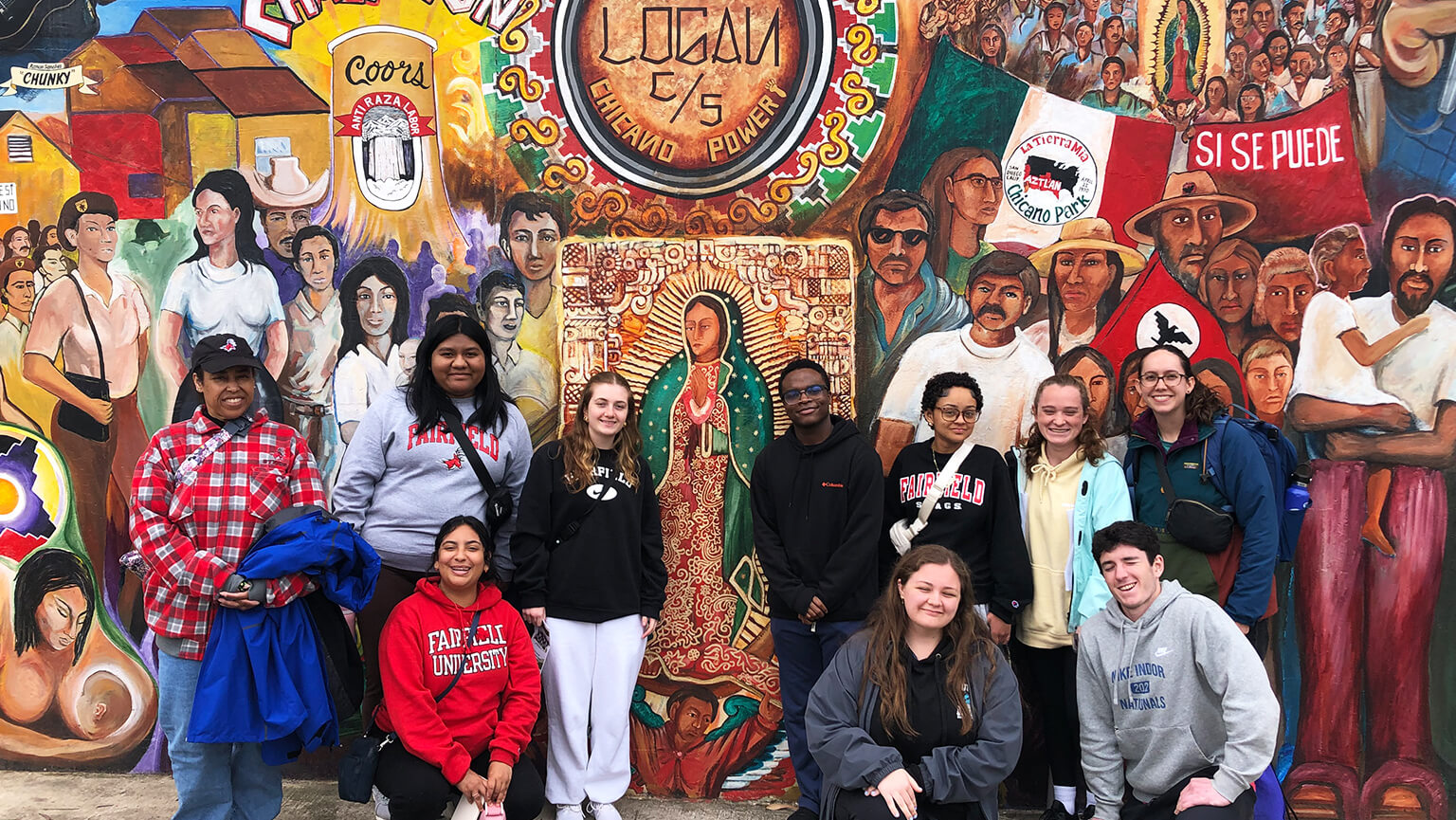 A diverse group of students poses together in front of a colorful mural, showcasing vibrant artwork and community spirit. 
