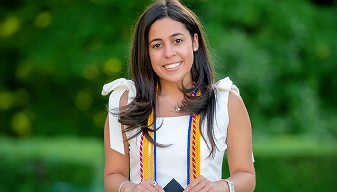 Graduate student smiling against a blurred, outdoor background while donning her commencement garb.