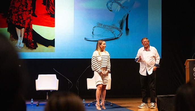 Female Dolan student standing on stage with Stuart Weitzman as he gives a presentation on shoe and fashion designs.