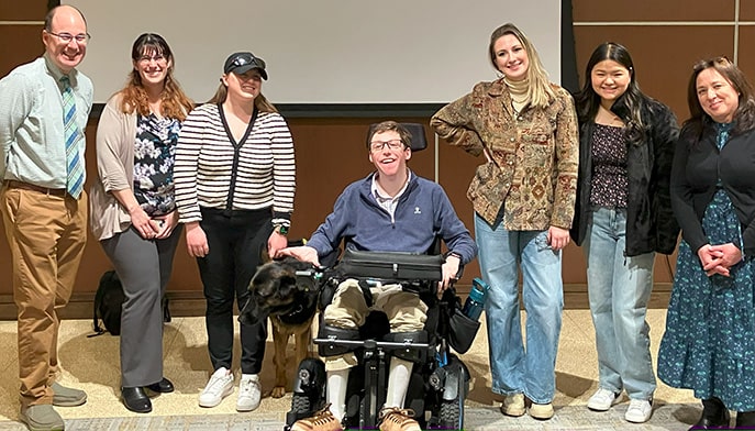 Students (one in a mobility wheelchair) posing in a group on stage after a successful disability awareness simulation.